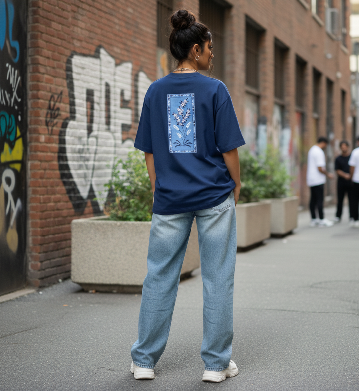 Person wearing a blue t-shirt with a floral design and light blue jeans standing on a street with graffiti.